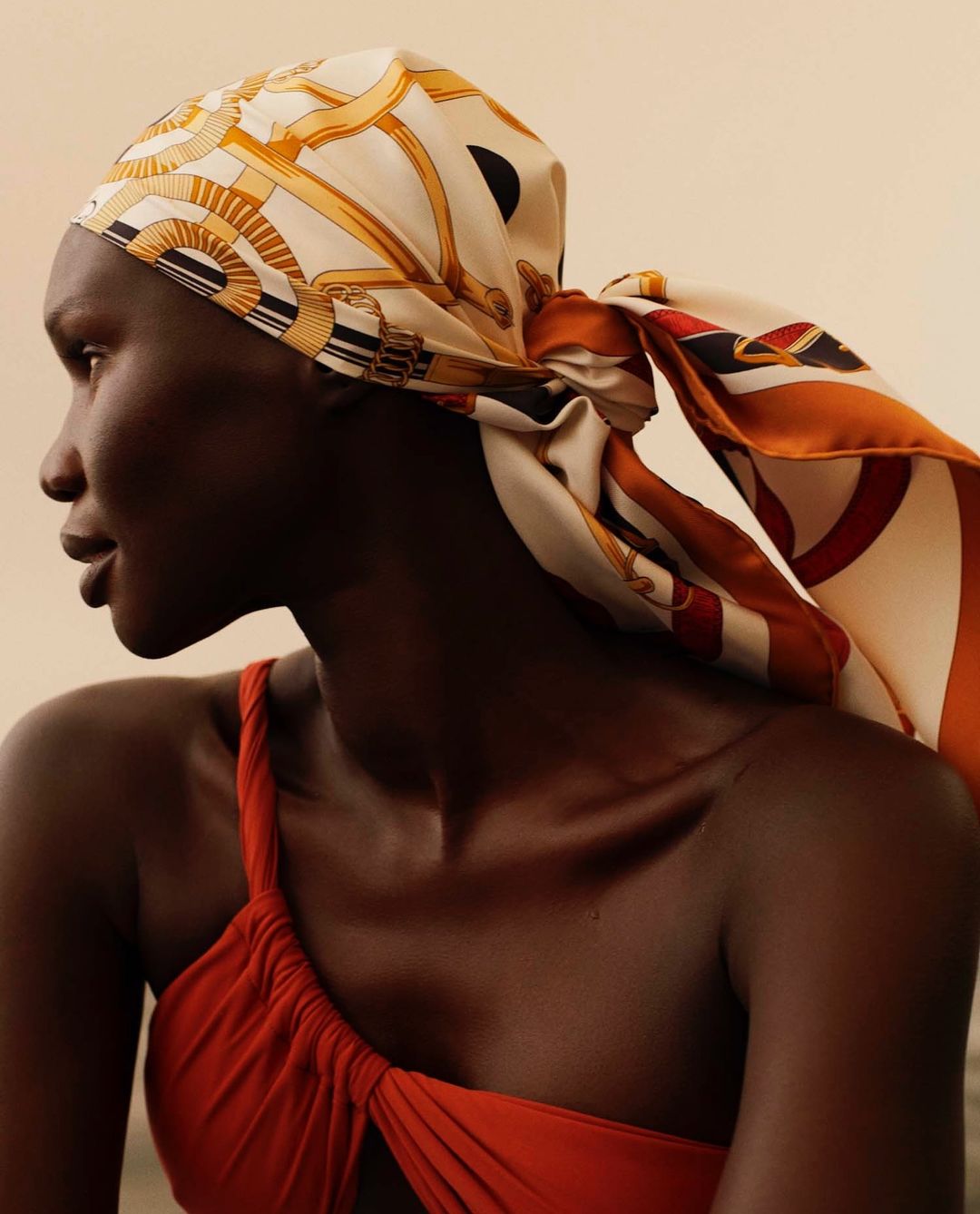 Profile view of an African American woman wearing a patterned headscarf and red one-shoulder top against a neutral background.