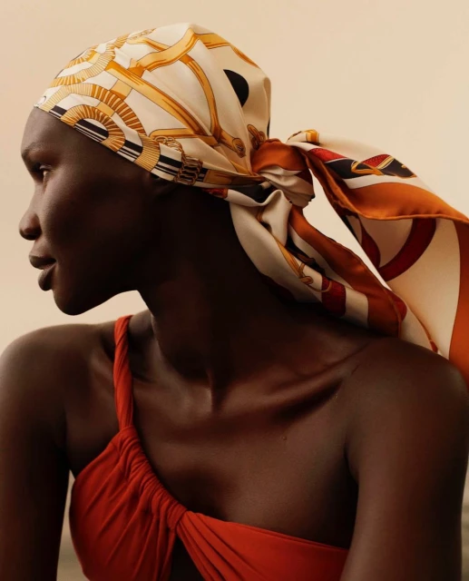 Profile view of an African American woman wearing a patterned headscarf and red one-shoulder top against a neutral background.