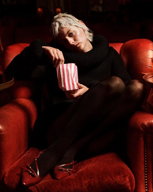 Woman in black outfit sitting in red theater chair holding striped popcorn bag in dim lighting with red shoes, tights, and short hair.