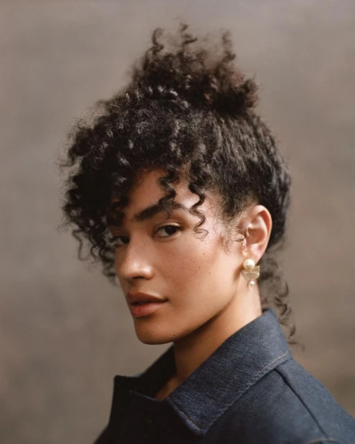 Close-up portrait of an adult female with curly hair wearing a denim jacket and flower earrings against a brown background.