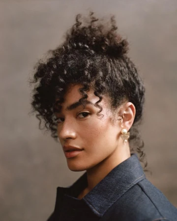 Close-up portrait of an adult female with curly hair wearing a denim jacket and flower earrings against a brown background.