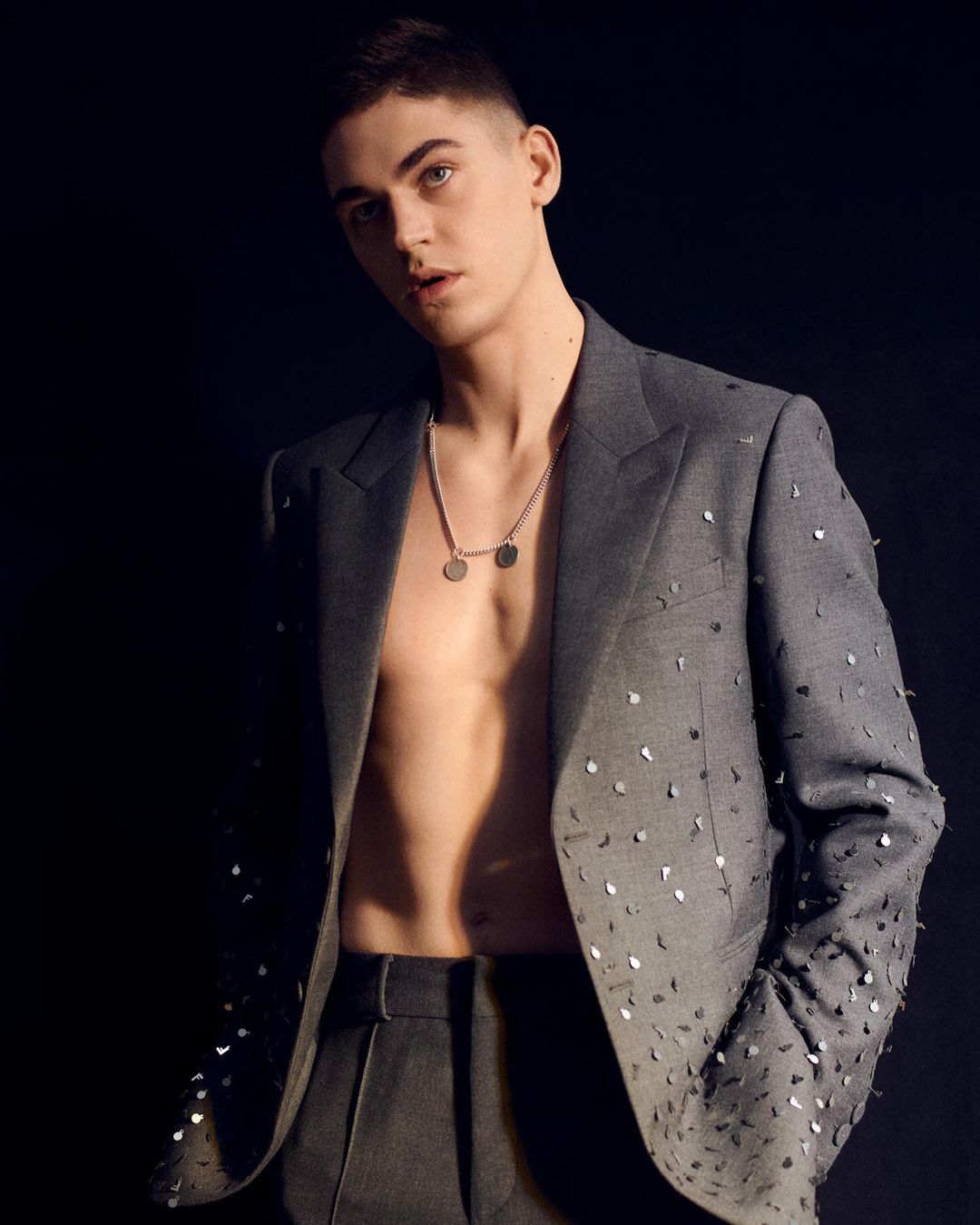 A young adult male with short hair, wearing a gray suit jacket with silver accents and a silver necklace posing against a black backdrop.