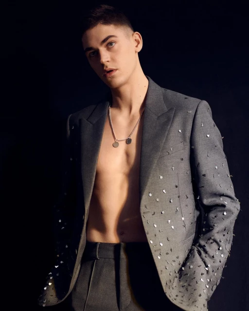 A young adult male with short hair, wearing a gray suit jacket with silver accents and a silver necklace posing against a black backdrop.