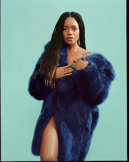 Portrait of an adult woman in a royal blue fur coat against an aqua blue backdrop, wearing long braids and jewelry, indoors.