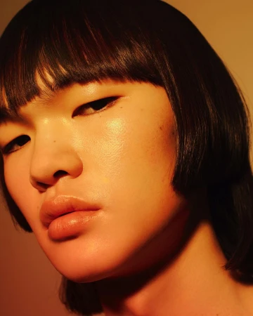 Close-up portrait of a young Asian woman with dark hair and a subtle expression, illuminated by warm-toned lighting in a studio setting.
