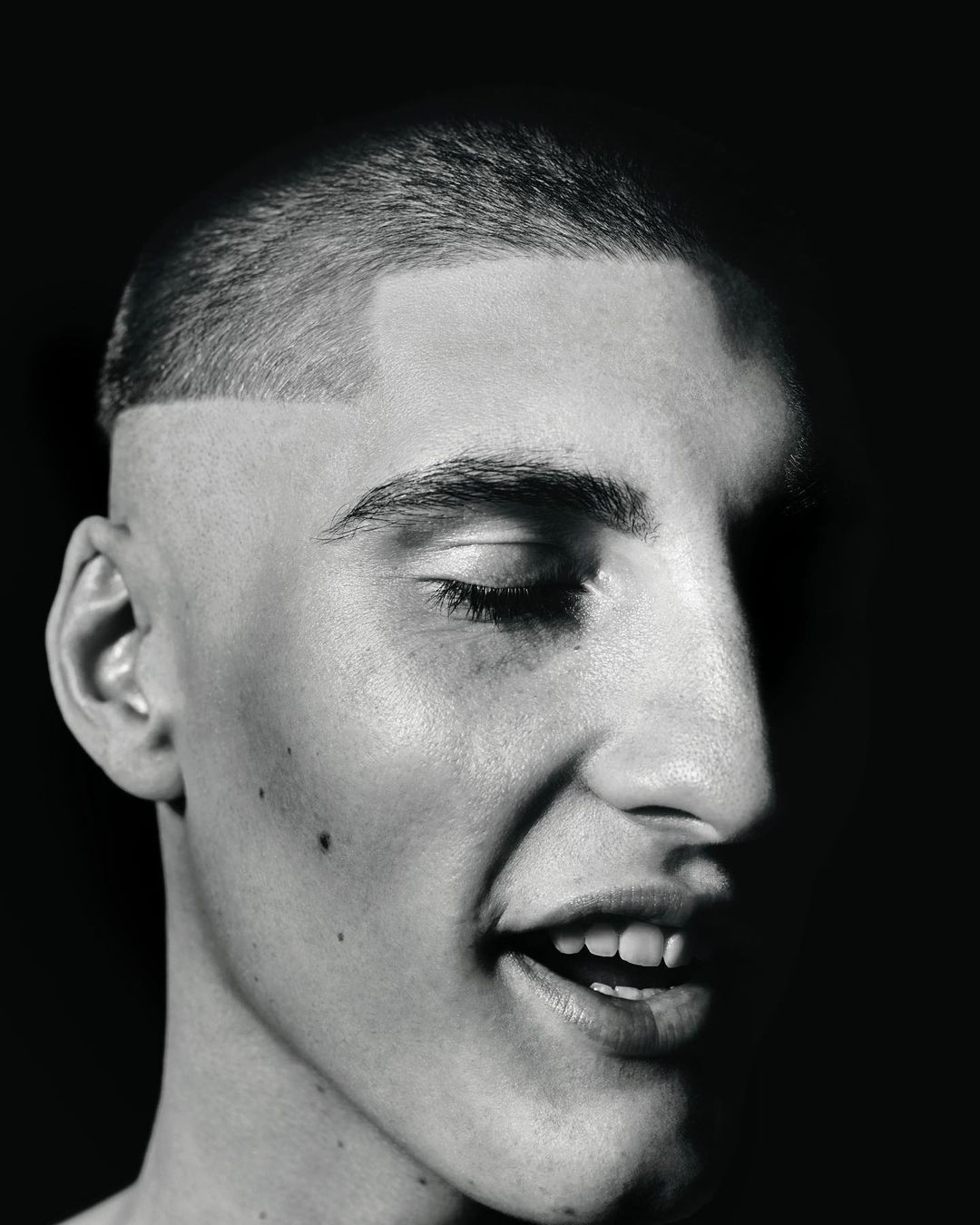 Striking black and white studio portrait of a young adult male with a buzzcut, fair skin, and a partially closed eye in a high contrast scene.
