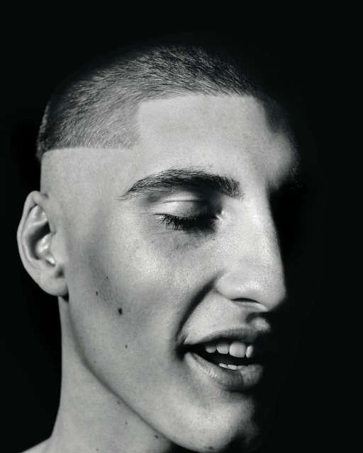 Striking black and white studio portrait of a young adult male with a buzzcut, fair skin, and a partially closed eye in a high contrast scene.