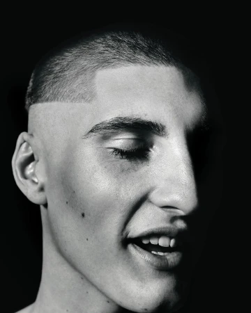 Striking black and white studio portrait of a young adult male with a buzzcut, fair skin, and a partially closed eye in a high contrast scene.