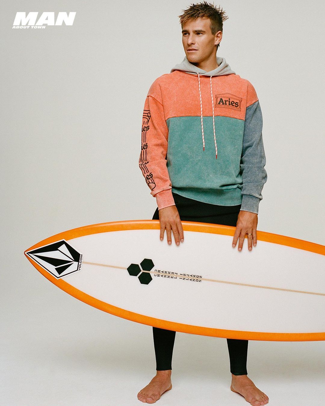 Male model holds a surfboard in a studio shot. He is wearing a color-block hoodie and black leggings and is barefoot.