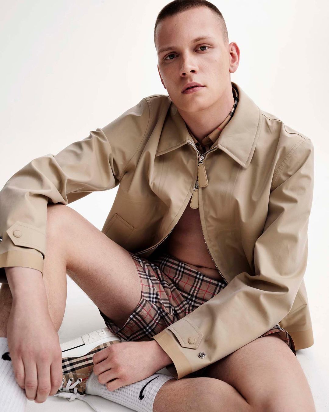 Young man with short buzz cut sits in a photo studio wearing a tan jacket, plaid shorts, white socks, and matching shoes. 