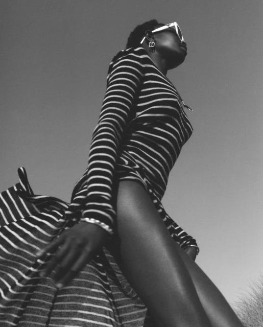 Black and white low angle portrait of an adult female model wearing sunglasses and a striped dress against a sky background.