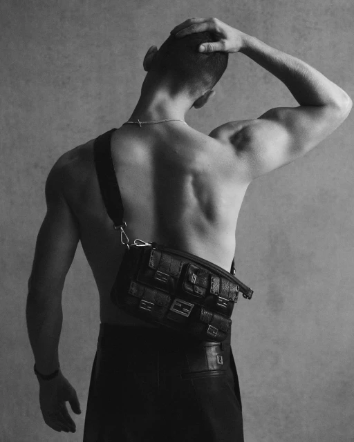 Black and white fashion shot of a male model with bare torso wearing leather pants and a woven crossbody bag against a grey backdrop.