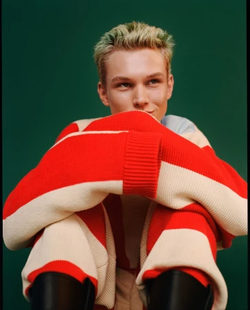 Young man with dyed blonde hair wearing a red and white striped sweater against a dark green backdrop