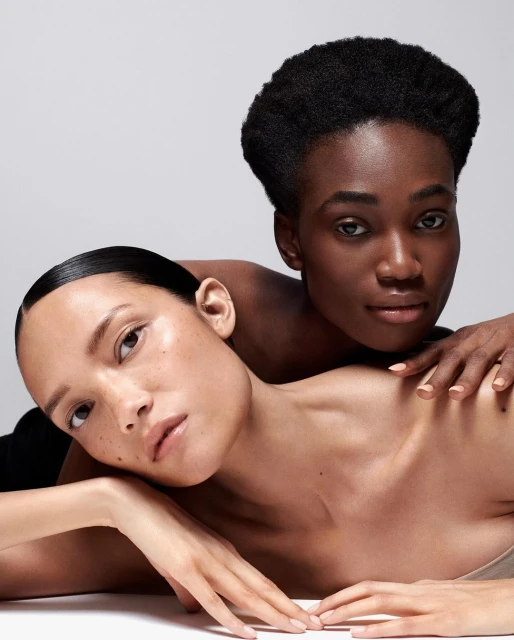 Close-up studio shot of two women with different skin tones in a beauty or fashion context, showcasing diverse representation and minimalist aesthetic.