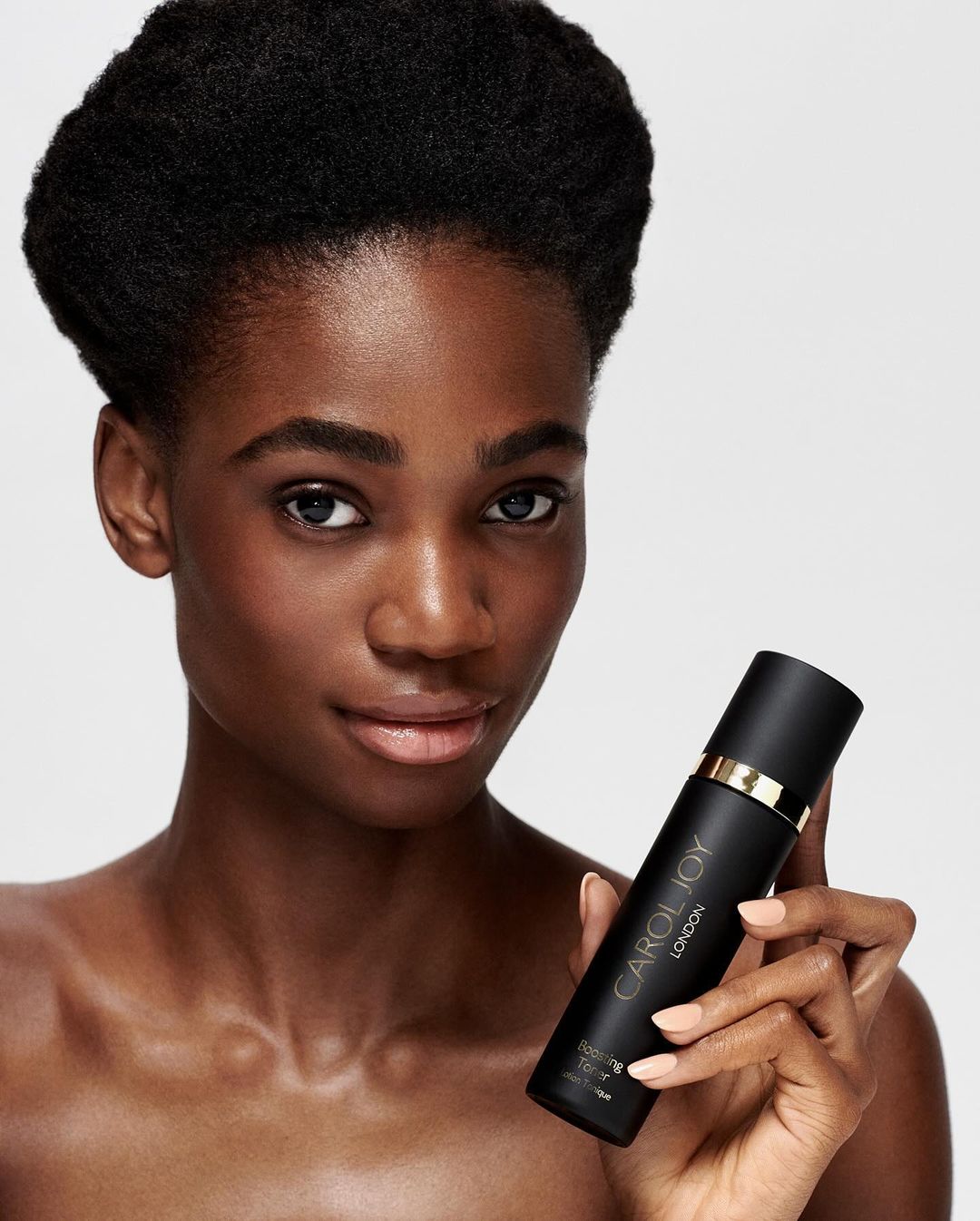 Close-up of a dark-skinned adult female holding a bottle of skincare product, set against a clean, neutral background.
