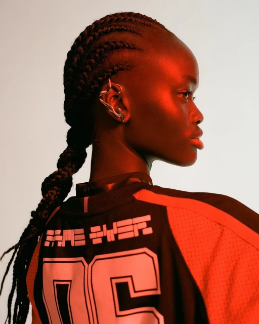 Close-up studio shot of an adult female fashion model with dark skin, braided hair, and a modern ear cuff wearing a black and orange jersey.