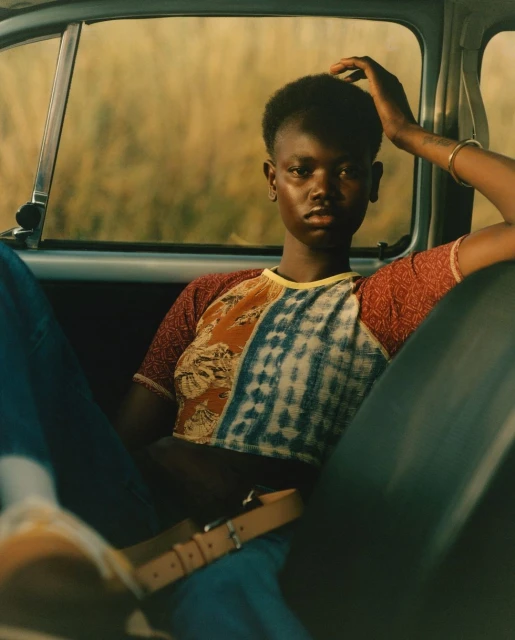 Portrait of a Black woman sitting in the backseat of a car. She wears jeans and a patterned crop top, with a field visible through the window.