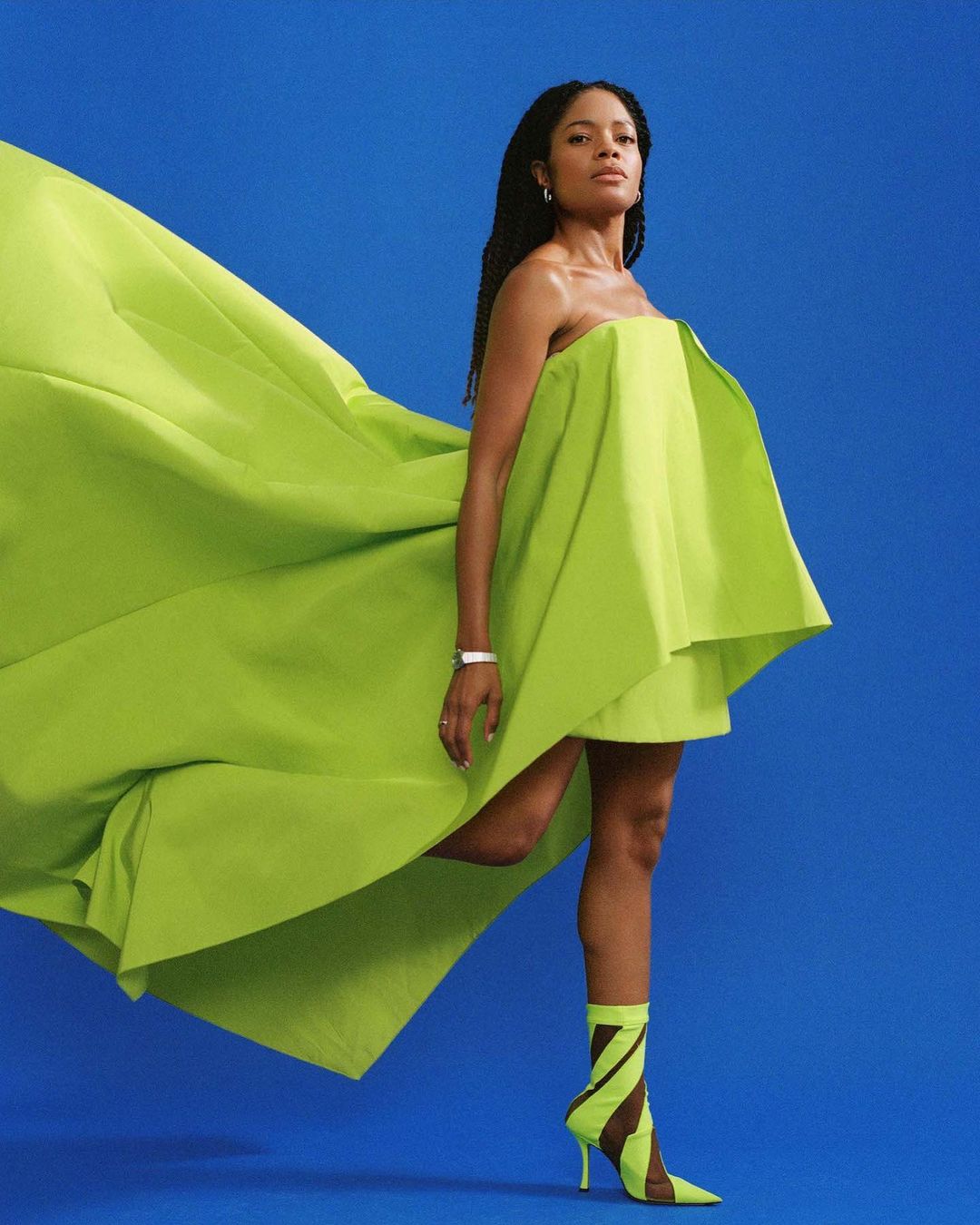Elegant woman poses in a vibrant chartreuse gown with sculptural details, paired with matching heels against a solid blue backdrop.