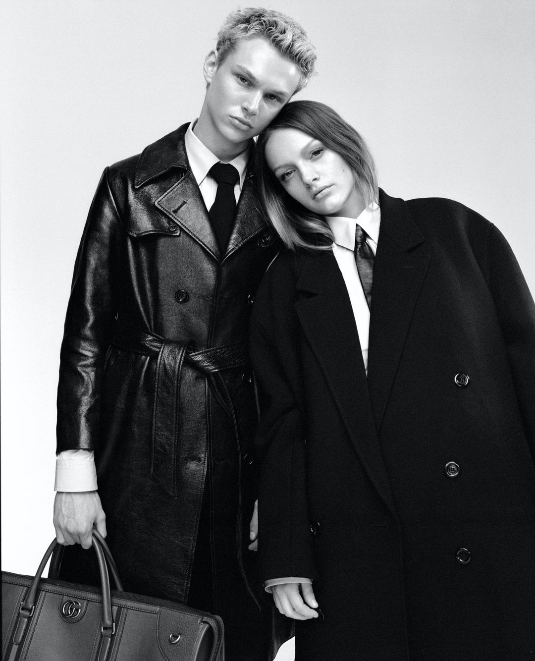 Black and white fashion shot of a blond male and a brunette female in dark overcoats and ties in a close up studio composition.