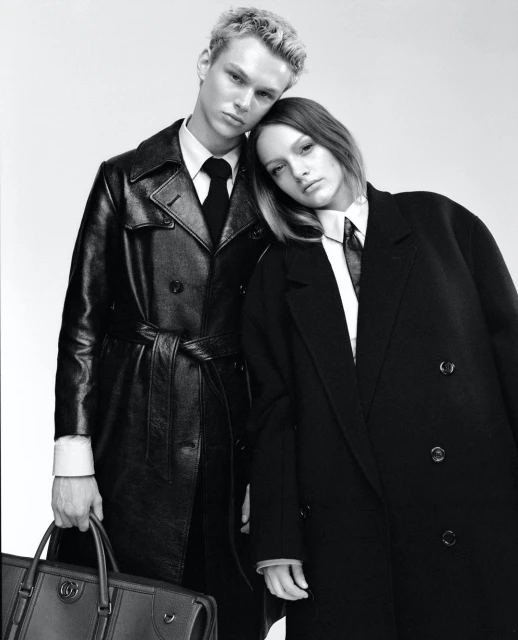 Black and white fashion shot of a blond male and a brunette female in dark overcoats and ties in a close up studio composition.