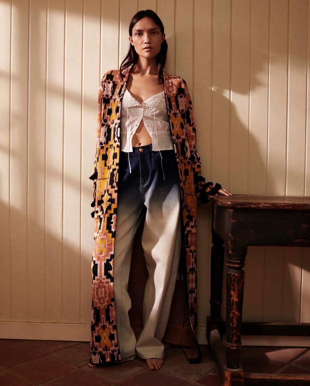 A woman stands indoors wearing a long patterned robe, white top, and blue jeans, posing next to a dark wooden table in soft, natural light.