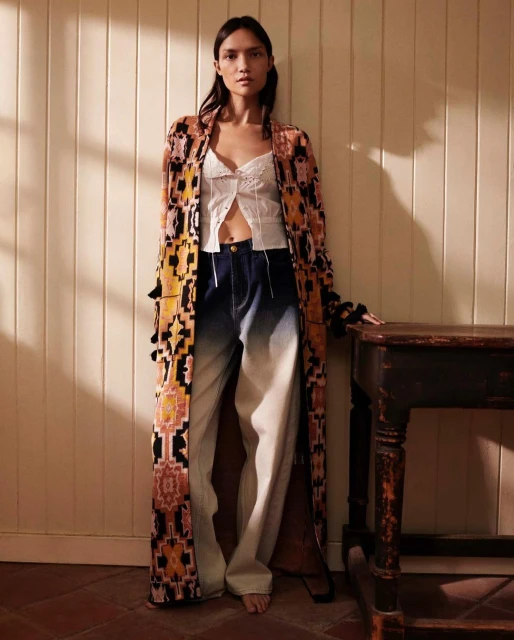 A woman stands indoors wearing a long patterned robe, white top, and blue jeans, posing next to a dark wooden table in soft, natural light.