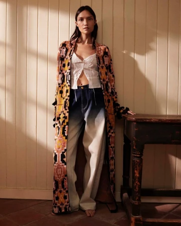 A woman stands indoors wearing a long patterned robe, white top, and blue jeans, posing next to a dark wooden table in soft, natural light.