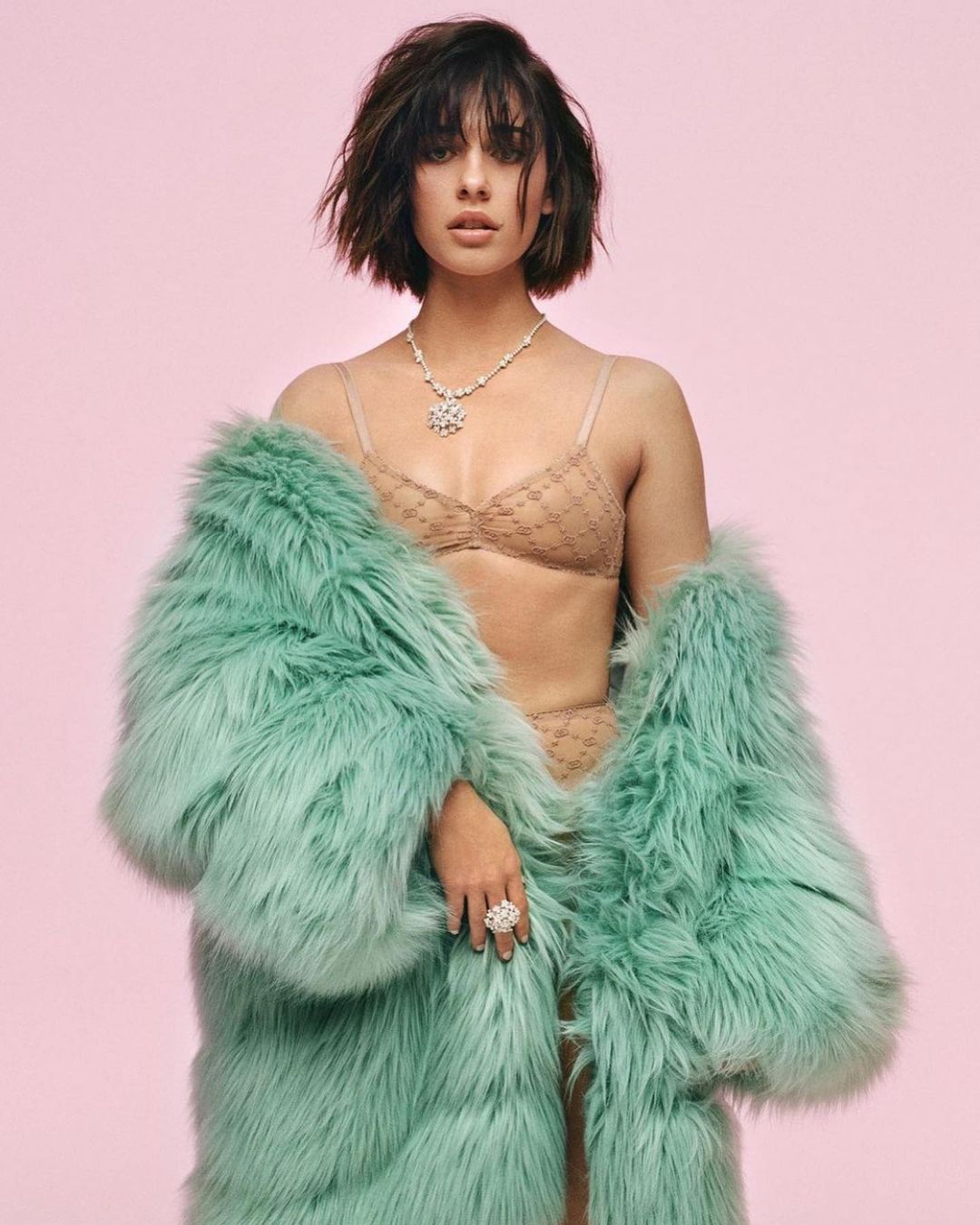 Attractive young woman in beige lingerie and a teal fur coat poses in a studio with a pink background, showcasing a necklace and ring.