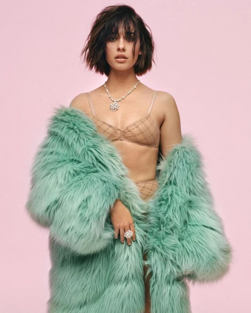 Attractive young woman in beige lingerie and a teal fur coat poses in a studio with a pink background, showcasing a necklace and ring.