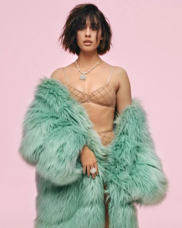 Attractive young woman in beige lingerie and a teal fur coat poses in a studio with a pink background, showcasing a necklace and ring.