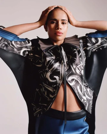 Model with hands on head wearing a black patterned jacket and blue pants against a light background