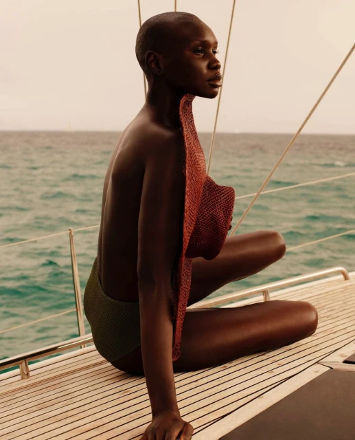 Adult female poses on a boat deck wearing green swimsuit, and holding a red sun hat with ocean and island in the background.