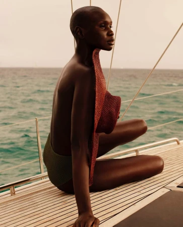 Adult female poses on a boat deck wearing green swimsuit, and holding a red sun hat with ocean and island in the background.