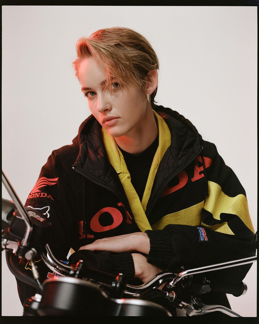 Portrait of a young adult in a black and yellow jacket posing in front of a motorcycle, set against a neutral studio backdrop.