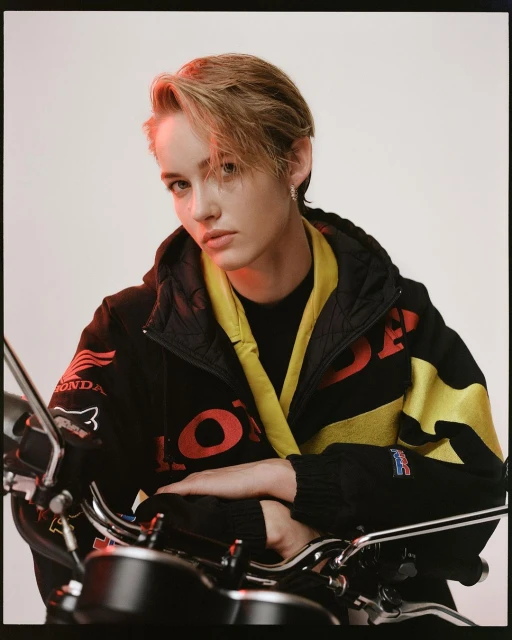 Portrait of a young adult in a black and yellow jacket posing in front of a motorcycle, set against a neutral studio backdrop.