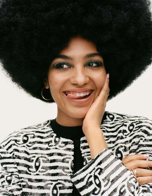 Portrait of a happy young woman with a large afro, wearing a sequined jacket, sticking her tongue out in a light studio setting.