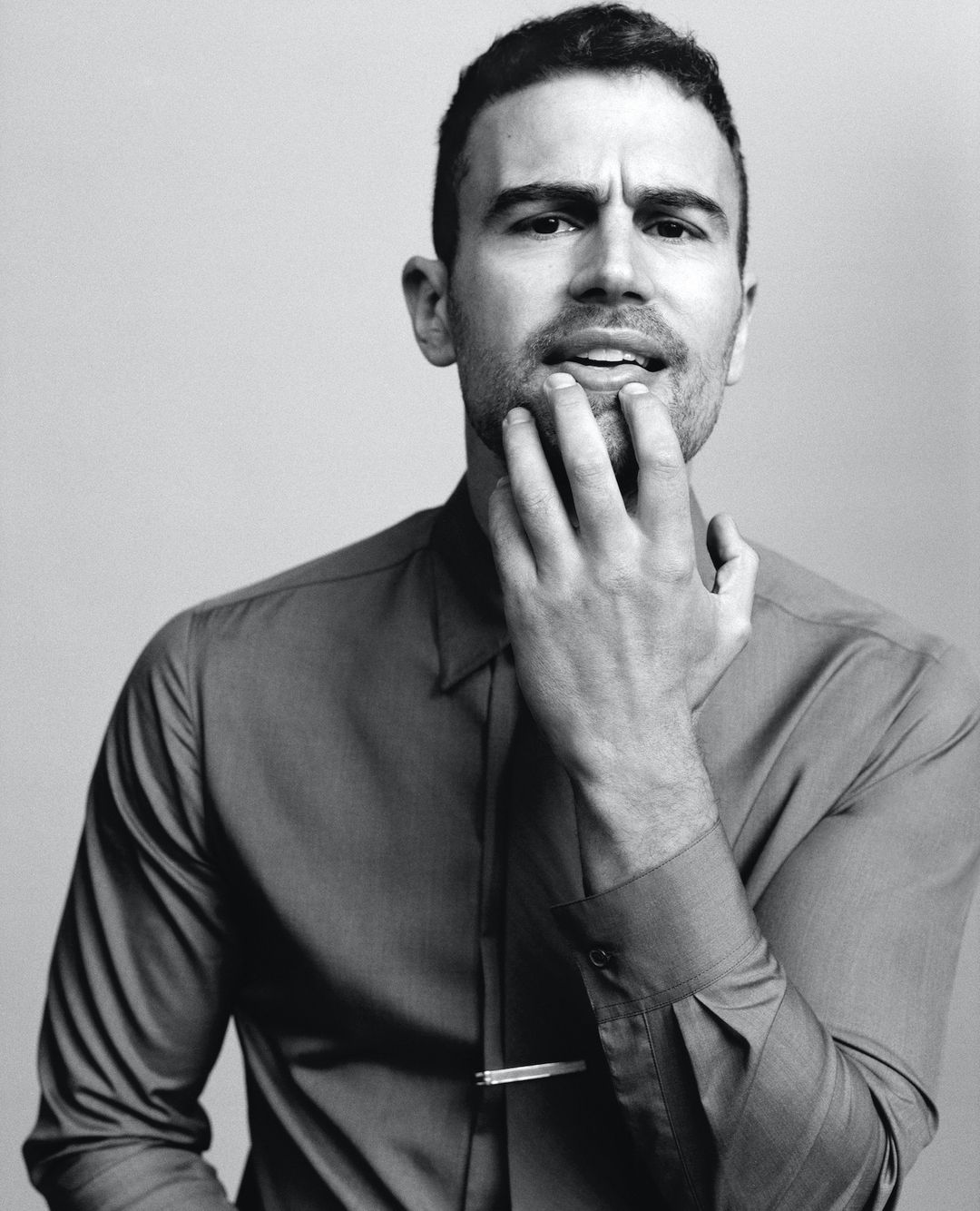 Portrait of a handsome adult male with his hand near his face, wearing a collared shirt and tie clip, against a plain background.