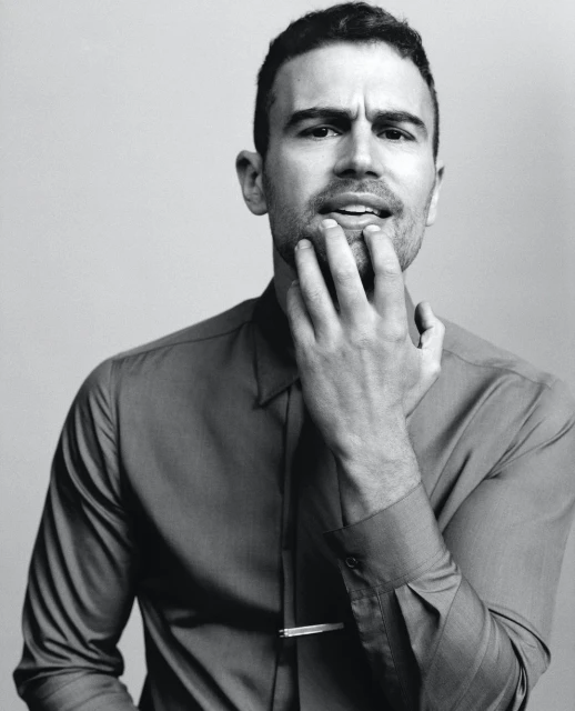 Portrait of a handsome adult male with his hand near his face, wearing a collared shirt and tie clip, against a plain background.