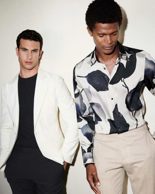 Two adult men in casual fashion stand indoors against a neutral wall, one in a white blazer and black t-shirt, the other in a patterned shirt.