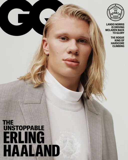 Close-up portrait of Erling Haaland on a magazine cover wearing a grey blazer, white top, and long blonde hair, against a light grey backdrop.