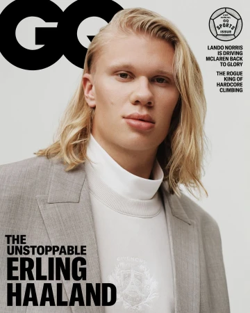 Close-up portrait of Erling Haaland on a magazine cover wearing a grey blazer, white top, and long blonde hair, against a light grey backdrop.
