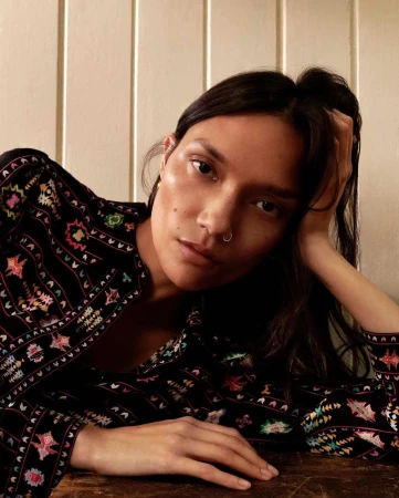 Close-up portrait of a woman wearing a black patterned blouse with colourful geometric embroidery, leaning on a wooden table.