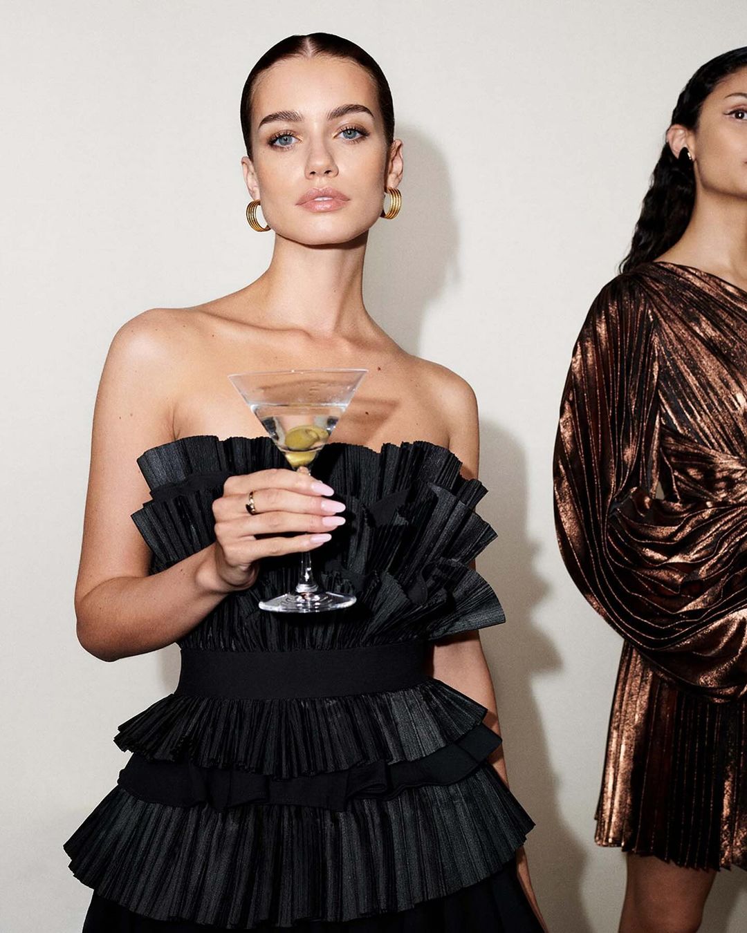Two women in dresses, one in black ruffles holding a martini glass with olives, the other in a metallic brown dress. Studio setting.
