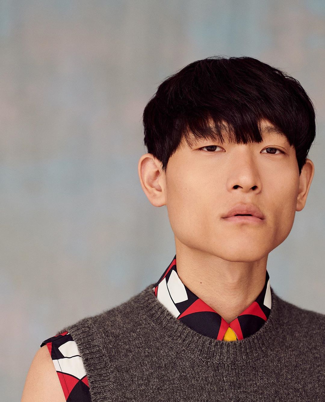 Portrait of a young adult Asian male with dark hair wearing a grey vest over a patterned shirt against a blurred background.