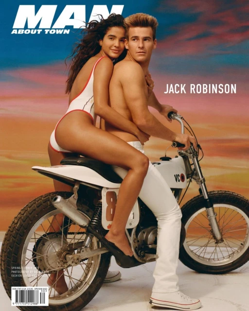 Magazine cover featuring a shirtless male model and a female model in a swimsuit, posing on a white vintage motorcycle against a sunset.