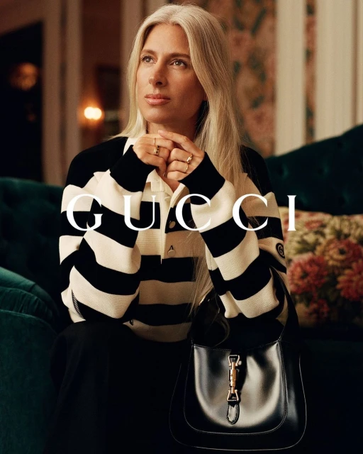 Adult woman with long grey hair in a black-and-white striped shirt poses with a black purse in an indoor setting with floral wallpaper.