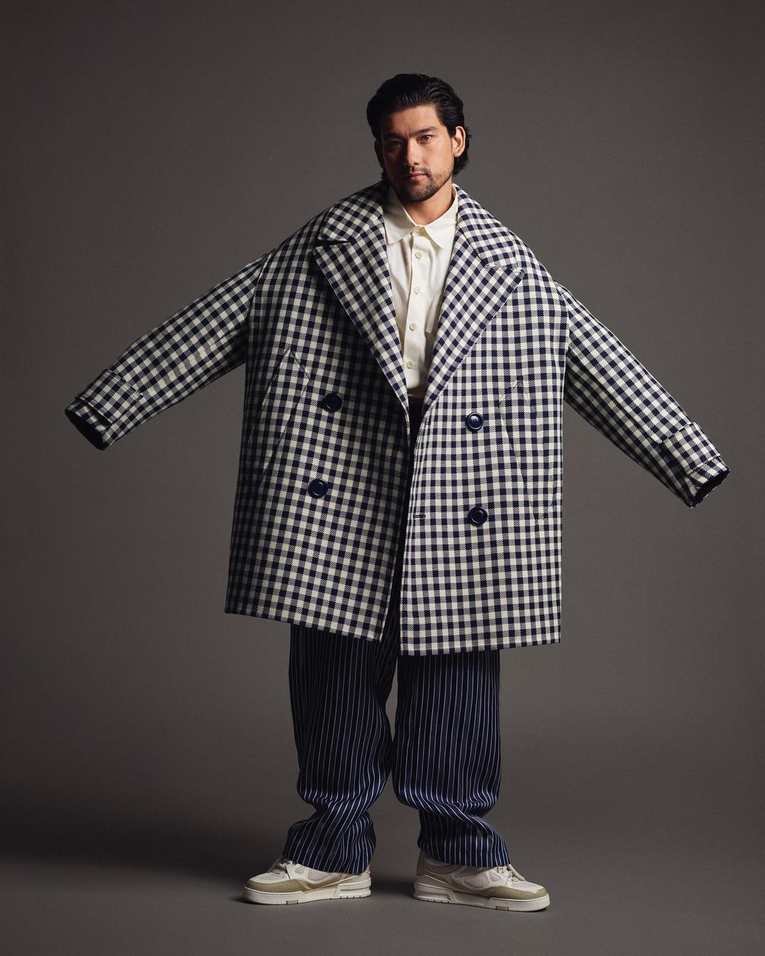 Man poses wearing a blue and white gingham coat and blue striped pants against a neutral gray background in a fashion editorial shot.