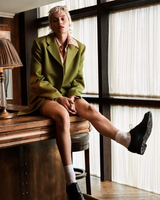 Stylish person sits on a counter wearing an olive green jacket, light pink shirt, dark skirt, socks, and shoes against a window backdrop.