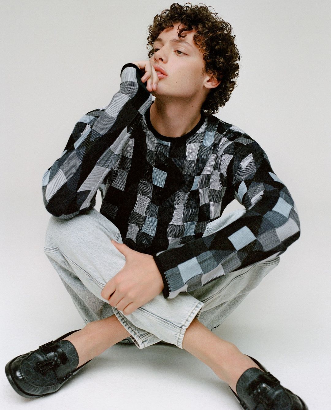 Young adult with curly hair poses indoors wearing a patterned sweater and jeans while seated against a white backdrop in a fashion pose.
