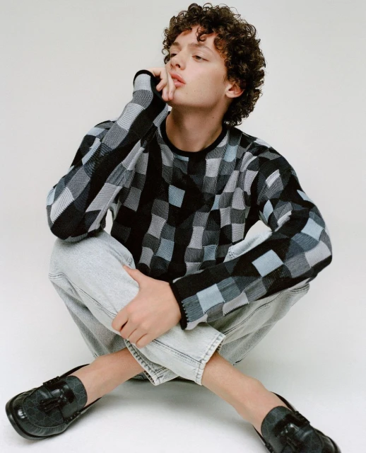 Young adult with curly hair poses indoors wearing a patterned sweater and jeans while seated against a white backdrop in a fashion pose.